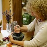 Woman using mortar and pestle to grind herbs at home, surrounded by dried flowers.
