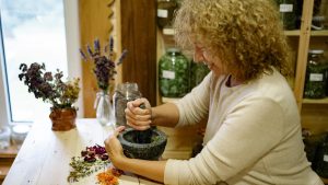 Woman using mortar and pestle to grind herbs at home, surrounded by dried flowers.