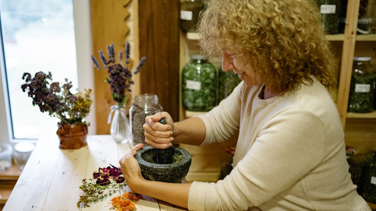 Woman using mortar and pestle to grind herbs at home, surrounded by dried flowers.