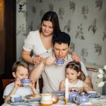 Contented family drinking tea with crepes together while sitting at table with candles at cozy kitchen with cuckoo clock on wall